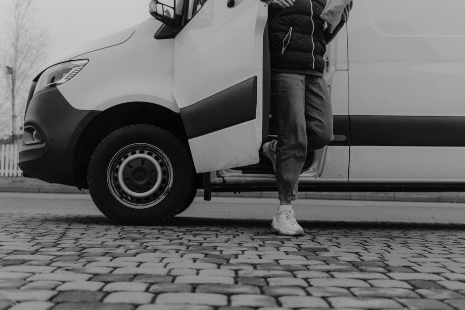 A person standing next to a white panel van on a cobblestone street, with one foot on the ground and the other partly inside the van. The individual is wearing jeans, a dark jacket, and white sneakers