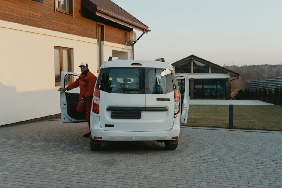 A person standing next to a white panel van on a cobblestone street, with one foot on the ground and the other partly inside the van. The individual is wearing jeans, a dark jacket, and white sneakers