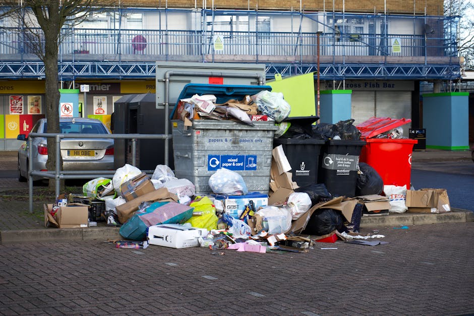 A large collection of overflowing public waste bins and recycling containers positioned on a paved sidewalk in front of a retail area in Isleworth, with various types of rubbish including cardboard boxes, paper, plastic bags, and packaging materials spilling onto the ground. Behind the bins, there are commercial storefronts and a building under construction or renovation with blue scaffolding. A parked grey car is visible to the left, and metal railings separate the waste area from the street. The scene captures a typical urban street location involved in household or commercial waste disposal, relevant to the logistics of house removals or packing and moving services, such as those provided by Man with Van Isleworth, especially when planning for proper waste management during relocation processes.
