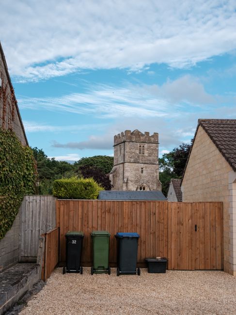 The image shows a rear view of a residential property in Isleworth, with a wooden fence separating the backyard from the street. In the yard, there are four large wheelie bins in black, green, blue, and black colours, positioned on a gravel surface. The bins are aligned against the fence, which has a small gate on the left side. Behind the fence, a historic stone tower with battlements and small windows is visible, indicating a church or historic structure nearby. The sky above is partly cloudy with patches of blue, and blooming trees and bushes surround the property, contributing to a peaceful residential environment. The scene appears to be set during daylight hours, with natural lighting illuminating the area, and the overall setting suggests a typical home environment where furniture or household items could be prepared for relocation or moving services by a professional team like Man with Van Isleworth, especially when considering local regulations such as skip permit requirements for the area.
