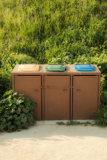 A brown outdoor trash bin with three lids in yellow, green, and blue, positioned on a concrete pathway next to green foliage and a grassy area. The bin is set against a backdrop of tall green grass and plants, indicating an outdoor location. The scene is lit by natural daylight. The image may relate to waste disposal during home relocation or packing processes involved in furniture transport or house removals, as provided by Man with Van Isleworth. The overall setting suggests a residential environment during waste collection or disposal effort connected to furniture removal and moving services in Isleworth.