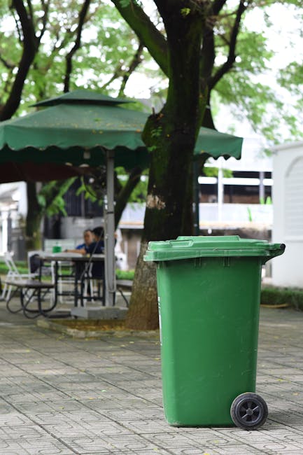 A green wheeled bin with a closed lid positioned on a paved sidewalk beneath a large tree with green foliage, in an outdoor setting that appears to be a park or public space. In the background, there is a person sitting at a table under a large green umbrella, with other tables and chairs visible, suggesting a communal area. The scene includes some structures and additional trees, with natural daylight illuminating the area. This outdoor environment may be relevant for removal and disposal services, aligning subtly with home relocation or furniture disposal topics discussed on the page about bulky waste options in Isleworth, as offered by Man with Van Isleworth.