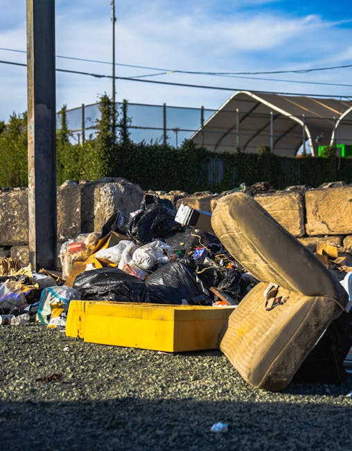 A brown outdoor trash bin with three lids in yellow, green, and blue, positioned on a concrete pathway next to green foliage and a grassy area. The bin is set against a backdrop of tall green grass and plants, indicating an outdoor location. The scene is lit by natural daylight. The image may relate to waste disposal during home relocation or packing processes involved in furniture transport or house removals, as provided by Man with Van Isleworth. The overall setting suggests a residential environment during waste collection or disposal effort connected to furniture removal and moving services in Isleworth.