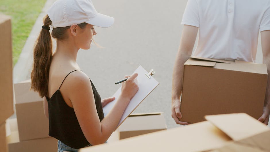 A young woman with light skin, wearing a white baseball cap and a black spaghetti strap top, is standing outdoors on a driveway or pavement during daytime. She is holding a clipboard and a pen, taking notes or recording details as part of a house removal or packing process. Behind her, there are several cardboard boxes, some partially open, and others sealed with packing tape, ready for transportation. Next to her, a person with light skin, wearing a white T-shirt, is holding a medium-sized cardboard box, preparing to load it into a moving van. The background suggests a residential setting with a focus on the logistics of packing and furniture transport. The scene depicts the preparation stage of a home relocation, with visible packing materials and the typical environmental lighting for an outdoor moving or loading activity, with Man with Van Isleworth providing professional removals services where packing, loading, and transportation are key elements.