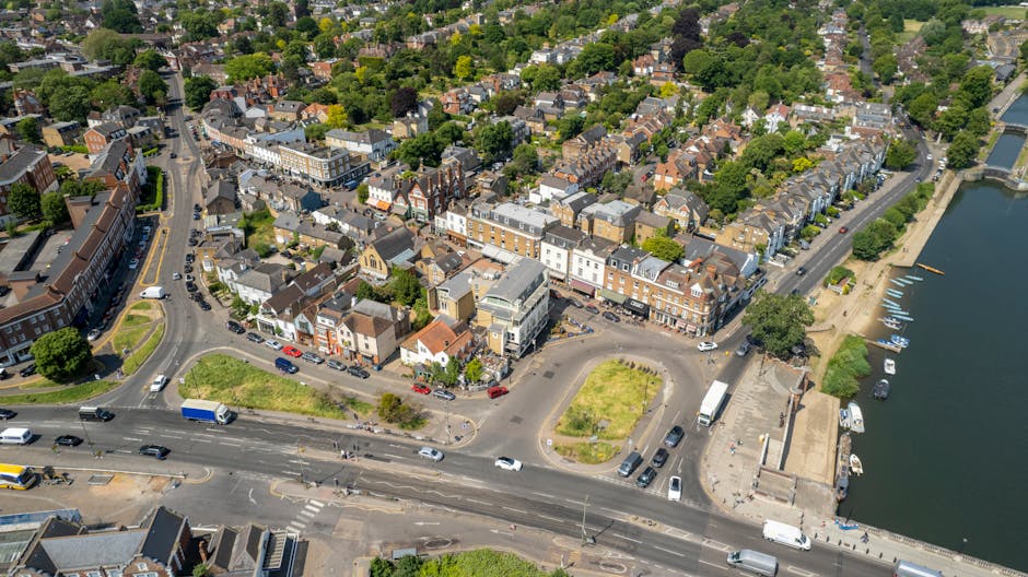 Aerial view of Isleworth High Street showing a range of terraced and brick buildings, with parked cars along the roadside and on side streets. The street runs parallel to a calm river with a small pie