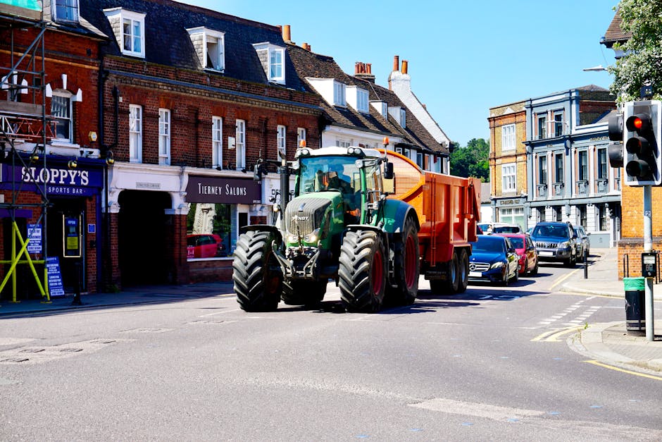 A large green tractor with an orange cargo trailer attached is positioned in the middle of a street in Isleworth, during a day with clear blue skies. The tractor appears to be in the process of transporting furniture or moving boxes, possibly as part of a home relocation. To the left, a shopfront with signage for 'Sloppy's Sports Bar' and 'Tierney Salons' is visible, with a yellow safety barrier nearby. Multiple cars are parked along the street, and a traffic light is visible at the corner, controlling the flow of traffic. The surrounding buildings are traditional brick and painted structures, with residential homes and commercial premises. The area is well-lit with natural sunlight, emphasizing the urban environment of a house move or furniture transport staged in this quiet street. Man with Van Isleworth’s crew might be coordinating the loading process onto the vehicle or planning the next stage of the move, supporting safe and professional furniture transport in the local area.