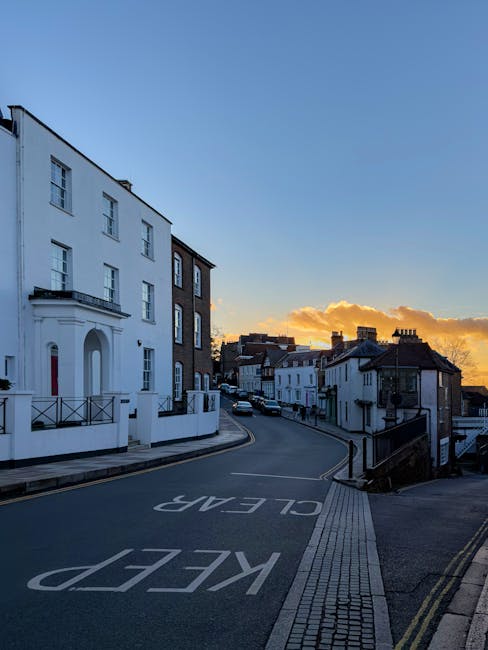 A narrow residential street in Isleworth during sunset, with a gentle curve in the road marked with 'KEEP CLEAR' in white letters painted directly on the asphalt. On the left side, there is a large white building with multiple windows, decorative architectural details, and a small balcony, indicating a residential property. The pavement is bordered by a tactile paving strip and a low wall, while on the right side, smaller white terraced houses with dark roofs line the street. The scene captures a moment of quiet before a house removal or furniture transport operation, with no visible vehicles or moving equipment present in the image. The lighting is soft, with a warm glow from the setting sun illuminating the clouds in the sky, and the overall environment suggests a typical quiet neighbourhood suitable for careful furniture transport and home relocation activities. Man with Van Isleworth routinely manages moving logistics in such urban streets, ensuring safe and efficient packing and loading processes.