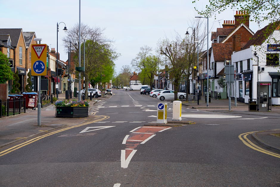 A large green tractor with an orange cargo trailer attached is positioned in the middle of a street in Isleworth, during a day with clear blue skies. The tractor appears to be in the process of transporting furniture or moving boxes, possibly as part of a home relocation. To the left, a shopfront with signage for 'Sloppy's Sports Bar' and 'Tierney Salons' is visible, with a yellow safety barrier nearby. Multiple cars are parked along the street, and a traffic light is visible at the corner, controlling the flow of traffic. The surrounding buildings are traditional brick and painted structures, with residential homes and commercial premises. The area is well-lit with natural sunlight, emphasizing the urban environment of a house move or furniture transport staged in this quiet street. Man with Van Isleworth’s crew might be coordinating the loading process onto the vehicle or planning the next stage of the move, supporting safe and professional furniture transport in the local area.