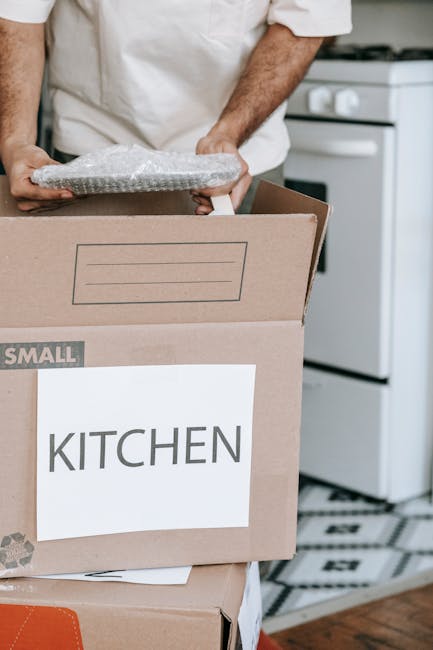A person dressed in a white shirt is seen carrying a small piece of bubble-wrapped packing material over a cardboard box labeled 'KITCHEN' in black letters, which is positioned inside a home setting near a white oven. The box is part of a moving process, with other packing boxes stacked nearby on the floor. The scene is well-lit, showing the interior of a kitchen with a black and white patterned floor. This image depicts packing activities associated with home relocation and furniture transport, relevant to professional removals services such as those provided by Man with Van Isleworth during a house move from Spring Grove to Syon Lane in Isleworth.