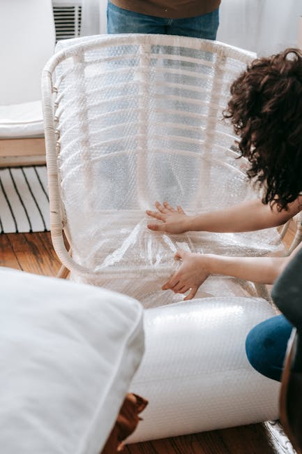 A wooden table inside a home contains packing materials such as bubble wrap, foam packaging, and brown paper, which are used for protecting household items during a house removal. There are several stacked rolls of bubble wrap and foam sheets, along with a small stack of cardboard boxes and a few rolls of packing tape. A clear plastic cup is placed in front of the packing supplies. The table is situated near a window, allowing natural light into the room, which has a radiator visible in the background. This scene depicts the preparatory stage of a home relocation, where packing is taking place before the furniture and boxes are loaded onto a vehicle for transit. The setup suggests an organized effort typical of professional removals, aligning with the services offered by Man with Van Isleworth in facilitating efficient furniture transport and packing and moving processes for clients in Isleworth.