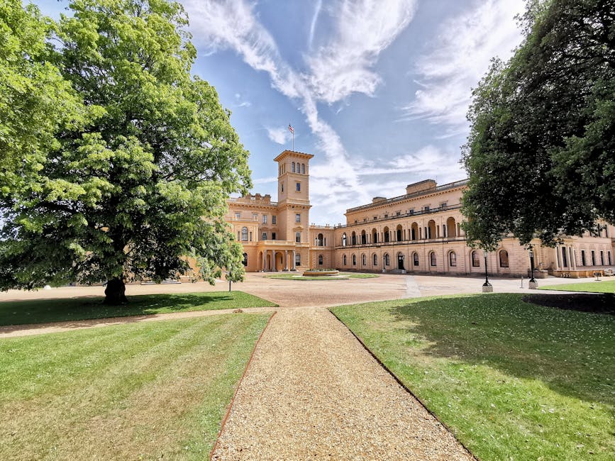 A view of Syon Park castle-like building with a central tower and arched windows, set against a partly cloudy sky. Two large, leafy green trees frame the scene on either side, with well-maintained grass and a gravel pathway leading directly to the entrance. The scene depicts an outdoor area suitable for a home relocation or furniture transport operation, with natural daylight illuminating the historic architecture and surrounding landscape. The image conveys a tranquil, open environment often relevant to house removals and moving logistics, such as access points for loading and unloading vehicles. Man with Van Isleworth offers advice on such access considerations for Isleworth residents, as highlighted on the page titled 'Syon Park moves: access tips for Isleworth residents'.