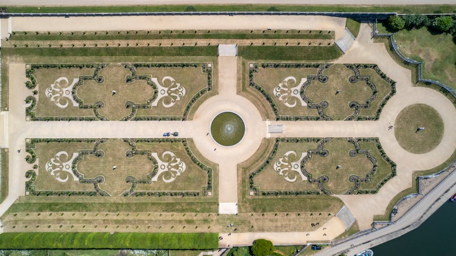 An aerial view of a symmetrical formal garden with four ornate, curved-shaped flower beds separated by straight gravel pathways, centered around a circular fountain with a small water feature. The flower beds contain neatly trimmed grass and decorative planting borders, with some small shrubs or topiary visible. A wide central pathway leads from the foreground to the fountain, flanked by additional lawns and walking paths. To the right, a rounded paved area with a person walking nearby is visible, and at the top, a road runs along the edge of the garden. The surrounding area includes a stone or brick wall, and the entire layout is part of a historic estate or park, often associated with large houses like Syon Park, which is relevant to house removals and relocation services. Man with Van Isleworth occasionally conducts furniture transport and packing efforts within such environments during home relocation projects.
