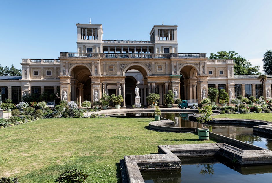 A view of Syon Park castle-like building with a central tower and arched windows, set against a partly cloudy sky. Two large, leafy green trees frame the scene on either side, with well-maintained grass and a gravel pathway leading directly to the entrance. The scene depicts an outdoor area suitable for a home relocation or furniture transport operation, with natural daylight illuminating the historic architecture and surrounding landscape. The image conveys a tranquil, open environment often relevant to house removals and moving logistics, such as access points for loading and unloading vehicles. Man with Van Isleworth offers advice on such access considerations for Isleworth residents, as highlighted on the page titled 'Syon Park moves: access tips for Isleworth residents'.